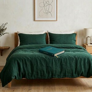 Dark emerald green linen bedding set on a wooden bed in a minimalist bedroom with natural wood furniture, soft neutral decor, and warm natural light.