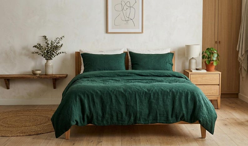 Dark emerald green linen bedding set on a wooden bed in a minimalist bedroom with natural wood furniture, soft neutral decor, and warm natural light.