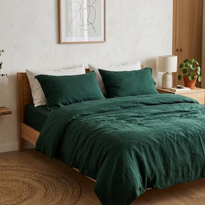 Dark emerald green linen bedding set on a wooden bed in a minimalist bedroom with natural wood furniture, soft neutral decor, and warm natural light.