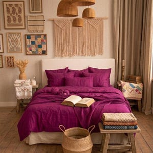 May include: A bedroom featuring a bed with deep magenta bedding, including a waffle-weave throw and several pillows. Above the bed hangs a macrame wall hanging and four woven pendant lights. A wooden stool holds folded textiles near the bed.