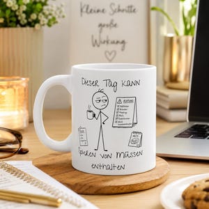 Könnte beinhalten: Ein weißer Keramikbecher mit einer schwarzen Strichzeichnung eines Strichmännchens, das eine Kaffeetasse hält. Der Text lautet: "Dieser Tag kann Spuren von Müssen enthalten", ergänzt durch eine To-Do-Liste und ein Warnschild mit "ACHTUNG".