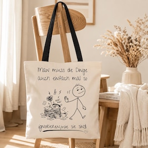 Könnte beinhalten: Eine cremefarbene Stofftasche mit schwarzen Tragegurten. Auf der Vorderseite befindet sich eine schwarze Strichzeichnung einer Strichmännchen-Figur, die einen unordentlichen Schreibtisch mit einem klingelnden Telefon ignoriert, sowie der Text: Man muss die Dinge auch einfach mal so ignorieren, wie sie sind.