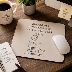 Könnte beinhalten: Beige Mauspad mit dem Text "Das Schönste an meinem Job ist, dass sich der Stuhl dreht!" und einer Strichmännchen-Figur. Eine weiße Maus, eine Kaffeetasse und ein Notizblock sind ebenfalls zu sehen.
