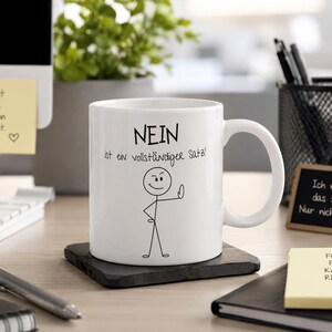 Könnte beinhalten: Eine weiße Keramik-Tasse mit dem deutschen Satz "NEIN ist ein vollständiger Satz!" und einer Strichmännchen-Figur, die einen Daumen nach unten zeigt. Die Tasse steht auf einem schwarzen quadratischen Untersetzer.