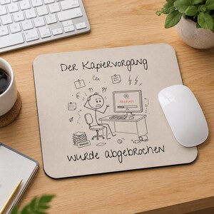 Może przedstawiać: Kwadratowa podkładka pod mysz o jasnobeżowej powierzchni z czarnym rysunkiem liniowym przedstawiającym postać z kresek przy komputerze, niemieckim tekstem "Der Kapiervorgang WURDE abgebrochen" oraz ekranem z komunikatem "Abbruch".