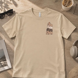 May include: A beige short-sleeved t-shirt featuring a small graphic on the left chest area. The graphic depicts a vertical stack of four cartoon capybaras in shades of brown, white, and tan, with a small orange fruit resting on the top capybara's head.