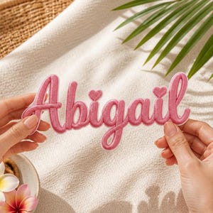 Puede incluir: Un parche bordado rosa con el nombre "Abigail" en fuente cursiva, con detalles de corazón. El parche se muestra sobre un fondo neutro con flores y follaje tropical.
