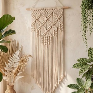 Puede incluir: Un tapiz de pared de macramé beige con un diseño geométrico y flecos. La decoración de la pared está suspendida de un pasador de madera y cuelga contra una pared de tono neutro. La pieza está rodeada de plantas.
