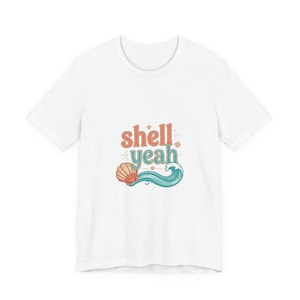 May include: White t-shirt with a colorful graphic that says "shell yeah". The design includes a seashell, a wave, and the text in orange and teal colors. The shirt is made of a soft material.