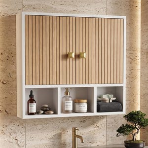 May include: A white wall-mounted bathroom cabinet featuring two doors with vertical light wood slats and square gold knobs. Below the doors are three open storage compartments containing a serum bottle, stones, lotion, a jar, and a folded towel.