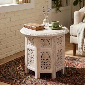 May include: A round, whitewashed wooden side table with intricate floral carvings on the panels and edge. On the table sits a brown leather-bound book, a clear glass vase containing dried lavender, and a green ceramic cup on a matching saucer.