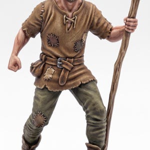 Könnte beinhalten: Eine bemalte Miniaturfigur einer bärtigen Person in einer beigen Tunika mit Flicken, einem braunen Gürtel mit Tasche und olivgrüner Hose. Die Figur wirkt robust, trägt braune Stiefel und hält einen hohen, knorrigen hölzernen Wanderstab in der Hand.