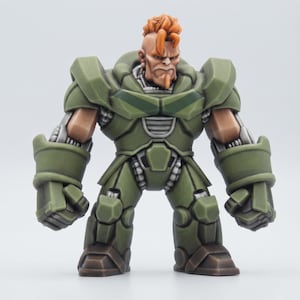 May include: A miniature figurine of a muscular character with orange hair and a goatee, wearing olive green armored plating over a metallic silver bodysuit. The figure has large, blocky gauntlets and boots, with a stern facial expression.