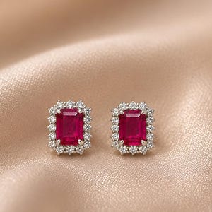May include: A pair of stud earrings featuring emerald-cut red rubies surrounded by a halo of small, round, clear white diamonds set in polished silver-toned metal. The earrings are positioned symmetrically, showcasing the vibrant red gemstone centres.