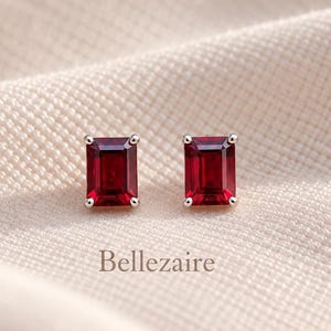 Può includere: Un paio di orecchini a lobo con gemme rosso intenso dal taglio rettangolare a smeraldo, incastonate in griffe di metallo color argento. La parola Bellezaire è stampata in piccole lettere scure sotto gli orecchini sul tessuto.