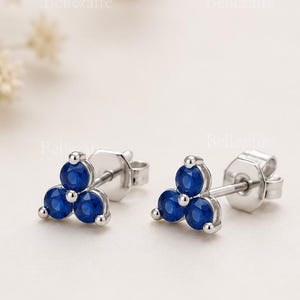 May include: A pair of stud earrings featuring three round, deep blue stones arranged in a triangular cluster. The stones are set in a polished silver-toned metal frame with a standard post and butterfly back closure for secure attachment.