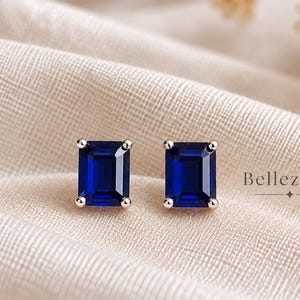 May include: A pair of stud earrings featuring rectangular emerald-cut deep blue stones set in silver-toned metal prongs. The text Bellezaire is printed to the right of the earrings. The earrings are positioned on a textured off-white fabric.