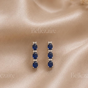 May include: A pair of vertical drop earrings featuring three oval-cut deep blue stones arranged in a column, accented by small clear stones at the top and between each blue stone. The word Bellezaire is printed multiple times across the image.