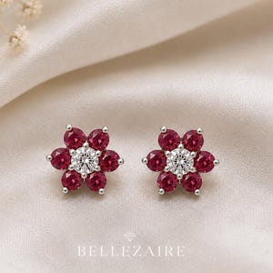 May include: A pair of stud earrings featuring a central clear round gemstone surrounded by six deep red round gemstones arranged in a flower shape, set in a silver-toned metal. The text BELLEZAIRE is printed below the earrings in white.