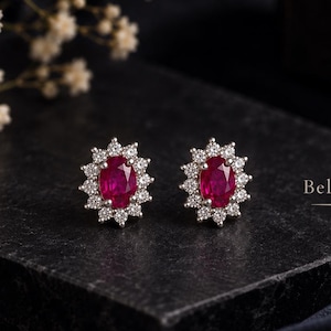 Puede incluir: Un par de pendientes de botón con rubíes de color rojo intenso en talla ovalada, rodeados por un halo de pequeñas piedras blancas redondas engastadas en metal plateado. El texto Bellezaire es visible a la derecha de la imagen.