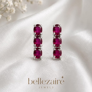 May include: A pair of silver-toned stud earrings, each featuring three vertically aligned oval-cut deep pink gemstones held in a prong setting. The text "bellezaire JEWELS" is printed in a dark brown font at the bottom of the image.