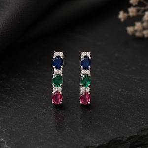 Può includere: Un paio di orecchini color argento con un design a barra verticale. Ogni orecchino è incastonato con tre pietre rotonde blu, verdi e rosse, accentuate da piccole pietre chiare tra di loro. La parola Bellezaire è stampata nell'angolo in basso a destra.