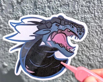 May include: A die-cut sticker featuring a stylised blue and black dragon head with an open mouth, sharp teeth, and a pink tongue. The dragon has dark scales on its head and neck, with light blue accents and a white border outline.