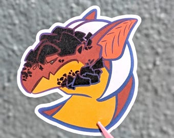 May include: A die-cut sticker featuring a stylised creature head with orange skin, a yellow neck, and black rocky textures on the forehead and jaw. The creature has a large pointed ear, a white curved crest, and a purple outline on a white border.