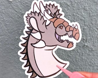 May include: A die-cut sticker featuring a stylised illustration of a dragon head in profile. The dragon is light grey with tan accents on the snout and horns, featuring a white underbelly and dark brown spikes along the back of its neck.