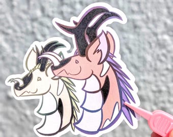 May include: A die-cut sticker featuring two stylised dragon-like creatures with horns and manes. The left creature is cream-coloured with dark accents, while the right creature is pink with purple manes and dark horns. A pink plastic tool touches the edge.