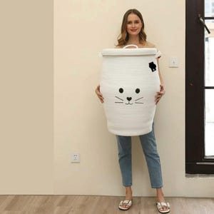 Può includere: Un grande cesto portabiancheria cilindrico in corda intrecciata bianca con coperchio. Il cesto presenta un disegno di muso di gatto ricamato in nero sulla parte anteriore, inclusi due occhi circolari, un piccolo naso triangolare e sei baffi.