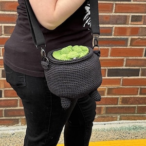 May include: A black crochet cauldron-shaped crossbody bag with a green lid. The bag has three small feet and a black adjustable strap. The cauldron is a unique accessory for Halloween or fantasy-themed outfits.