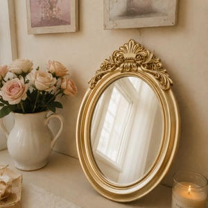 May include: An oval mirror with a gold-colored frame featuring ornate, carved detailing at the top. The mirror has a smooth, reflective surface and a rounded, polished border. The frame is constructed with a prominent, decorative crest design.