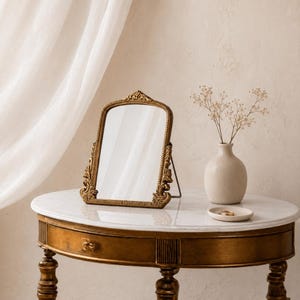 Könnte beinhalten: Ein goldfarbener, verzierter Schminkspiegel mit rechteckiger Form, der auf einem kleinen, runden Tisch mit Marmorplatte steht. Eine kleine Vase mit Trockenblumen und eine kleine Schale befinden sich ebenfalls auf dem Tisch. Der Tisch hat ein dunkles Holzgestell.