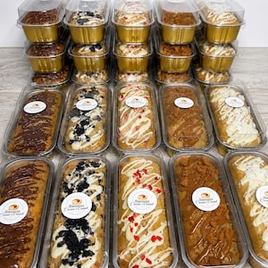 May include: Multiple rectangular banana bread loaves in clear plastic containers with gold foil bases. Each loaf features distinct toppings including chocolate drizzle, crushed cookies, red fruit pieces, or white icing, all labelled Banana Cream & Crumb.