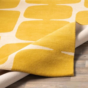 May include: A rectangular area rug featuring a repeating pattern of rounded yellow rectangles on a cream-colored background. One corner of the rug is folded over, revealing the textured pile surface and the underside of the fabric construction.