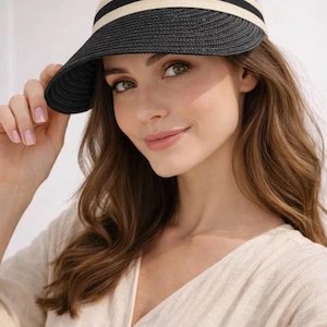 Puede incluir: Una visera de paja tejida en color negro con una banda de tela beige ancha que presenta dos finas rayas horizontales negras. La visera tiene un ala curva y estructurada, junto con una banda diseñada para ajustarse a la cabeza.