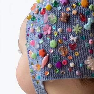 May include: A light blue knitted bonnet decorated with an assortment of colourful plastic charms, beads, and sequins. The embellishments feature shapes such as stars, flowers, shells, and small teddy bears scattered across the textured fabric surface.