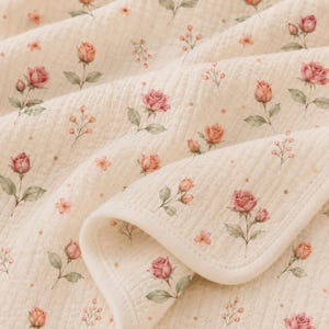 May include: A cream-colored quilted blanket featuring a repeating pattern of pink roses, rosebuds, and small green leaves. The fabric has a textured, crinkled appearance with a finished edge, showcasing a soft floral design across the surface.