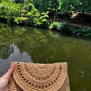 Könnte beinhalten: Beige geflochtene Stroh-Clutch mit Klappenverschluss. Die Klappe hat ein dekoratives, halbkreisförmiges Lochmuster. Der Hintergrund zeigt verschwommen Wasser und Bäume, was auf eine Umgebung im Freien hindeutet.