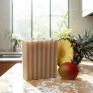 May include: A rectangular bar of beige soap with a vertically ribbed texture on its front surface. Next to the soap sits a halved pineapple with a spiky green crown and a single small, smooth mango with a gradient of red, orange, and green skin.