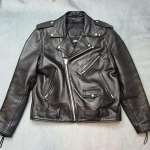May include: A black leather motorcycle jacket featuring an asymmetrical silver-tone zipper closure, wide notched lapels with snap buttons, shoulder epaulets, multiple zippered pockets, a small flap pocket, and a quilted interior lining.