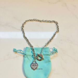 May include: A silver necklace with a toggle clasp and a heart-shaped pendant. The pendant is engraved with text and hangs from a chain. The necklace rests on a light blue drawstring pouch.