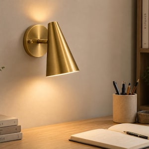 Peut inclure: Une applique murale en laiton brossé dotée d'un abat-jour conique fixé à une base circulaire. Le luminaire émet une lumière chaude dirigée vers le bas, éclairant la zone située en dessous. Le design comprend un bras simple reliant l'abat-jour au support.