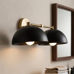 May include: A wall-mounted light fixture featuring two matte black dome-shaped shades attached to a horizontal brass-toned metal bar. The fixture includes a circular brass wall plate and two glowing light bulbs positioned inside the black domes.