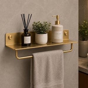 Puede incluir: Un estante de baño de pared en color oro mate con una barra para toallas debajo. El estante sostiene un difusor de varillas de vidrio negro, una pequeña planta verde en una maceta de cerámica blanca y un dispensador beige con la etiqueta Hand Wash.