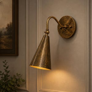 May include: A wall-mounted sconce light fixture featuring a conical shade and a curved arm, finished in an aged, mottled brass tone. The fixture includes a circular wall plate and a swivel joint, casting a warm, downward-facing light glow.