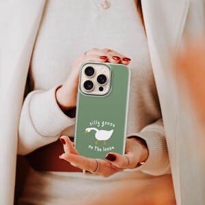 以下が含まれることがあります： セージグリーンのスマートフォンケース。オレンジ色の足とくちばしを持つ白いガチョウのイラストが描かれています。ガチョウのグラフィックの上下には、白い手書き風のフォントで「silly goose on the loose」という文字がプリントされています。