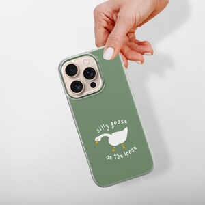 以下が含まれることがあります： セージグリーンのスマートフォンケース。白いガチョウのグラフィックと、手書き風の白いフォントで「silly goose on the loose」というテキストが描かれています。ケースには透明な縁があり、背面のカメラレンズとフラッシュ用の切り抜きがあります。