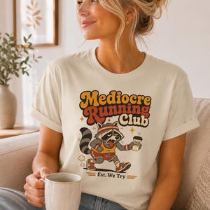 May include: Cream-colored t-shirt featuring a cartoon raccoon running with a coffee cup. The shirt has the text "Mediocre Running Club" in orange and brown, with "Est. We Try" below. The raccoon is wearing a headband and running shorts.
